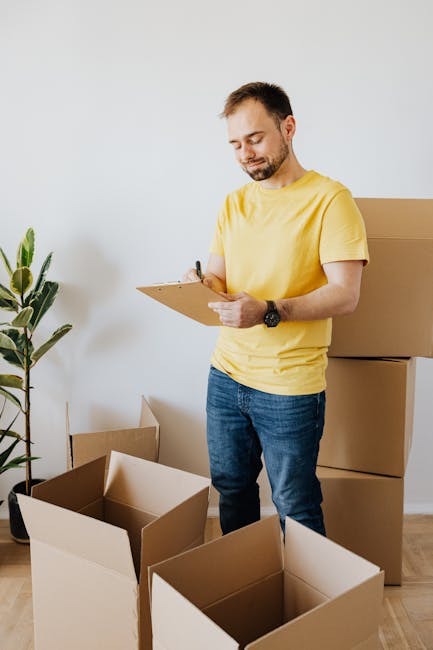 A man with a beard and short dark hair, dressed in a yellow t-shirt and blue jeans, standing in a bright room with a plain white wall, surrounded by several empty cardboard moving boxes. He is holding